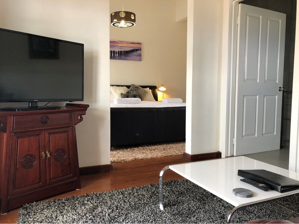 A room with a TV on a wooden cabinet, a bed with a white headboard, a painting on the wall, and a door.