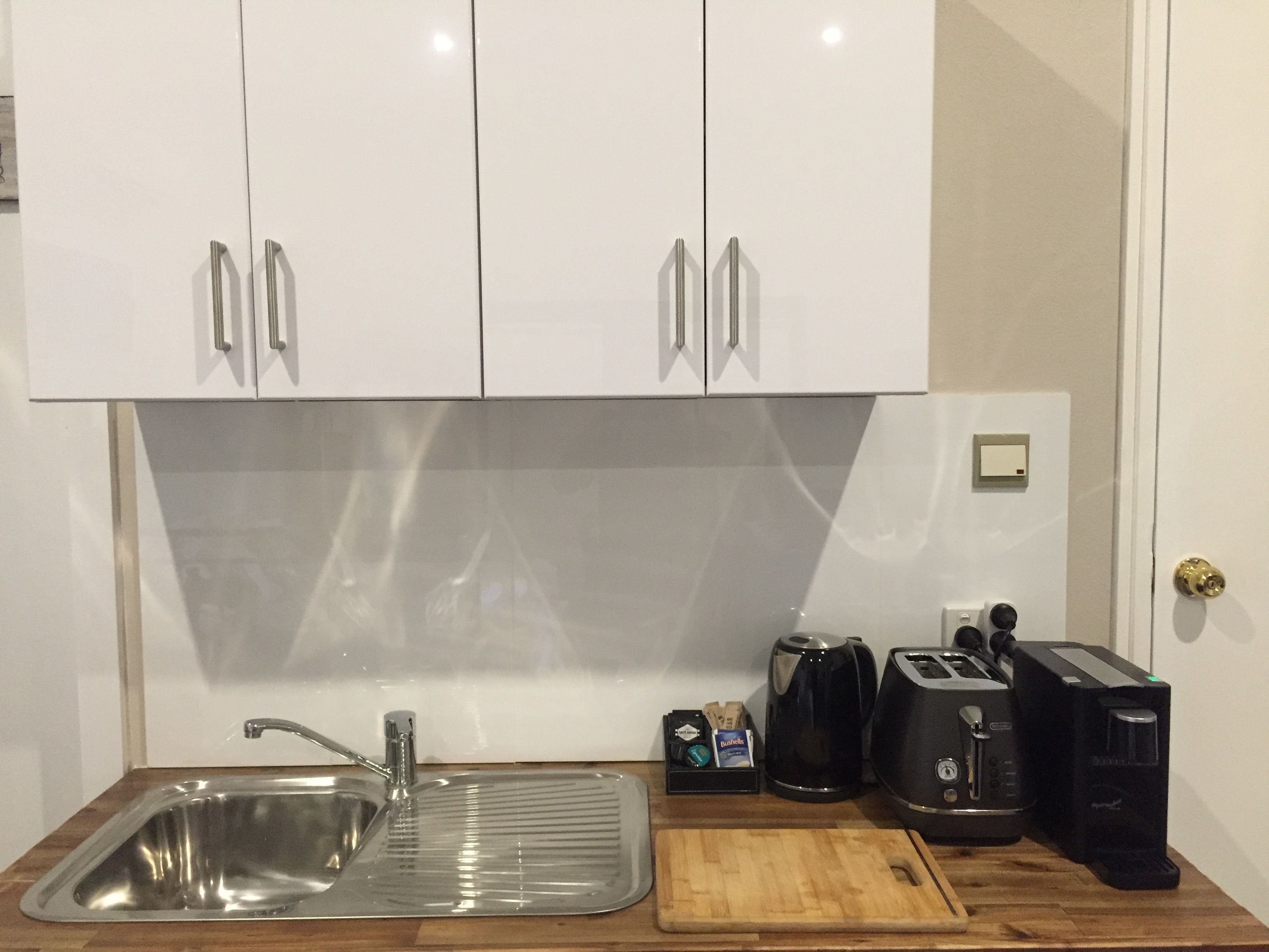 A kitchen with white cabinets, a stainless steel sink, a wooden cutting board, a coffee maker, a toaster, and a kettle.