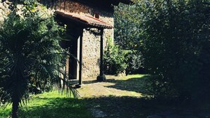 Un bâtiment rustique à la façade en pierre et au toit de tuiles, entouré d’une végétation luxuriante et bordé d’un chemin pavé.