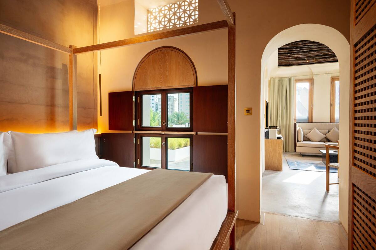 A hotel room with a large bed, a wooden headboard, a window with a view of greenery, and a doorway leading to a sitting area with a sofa and a table.