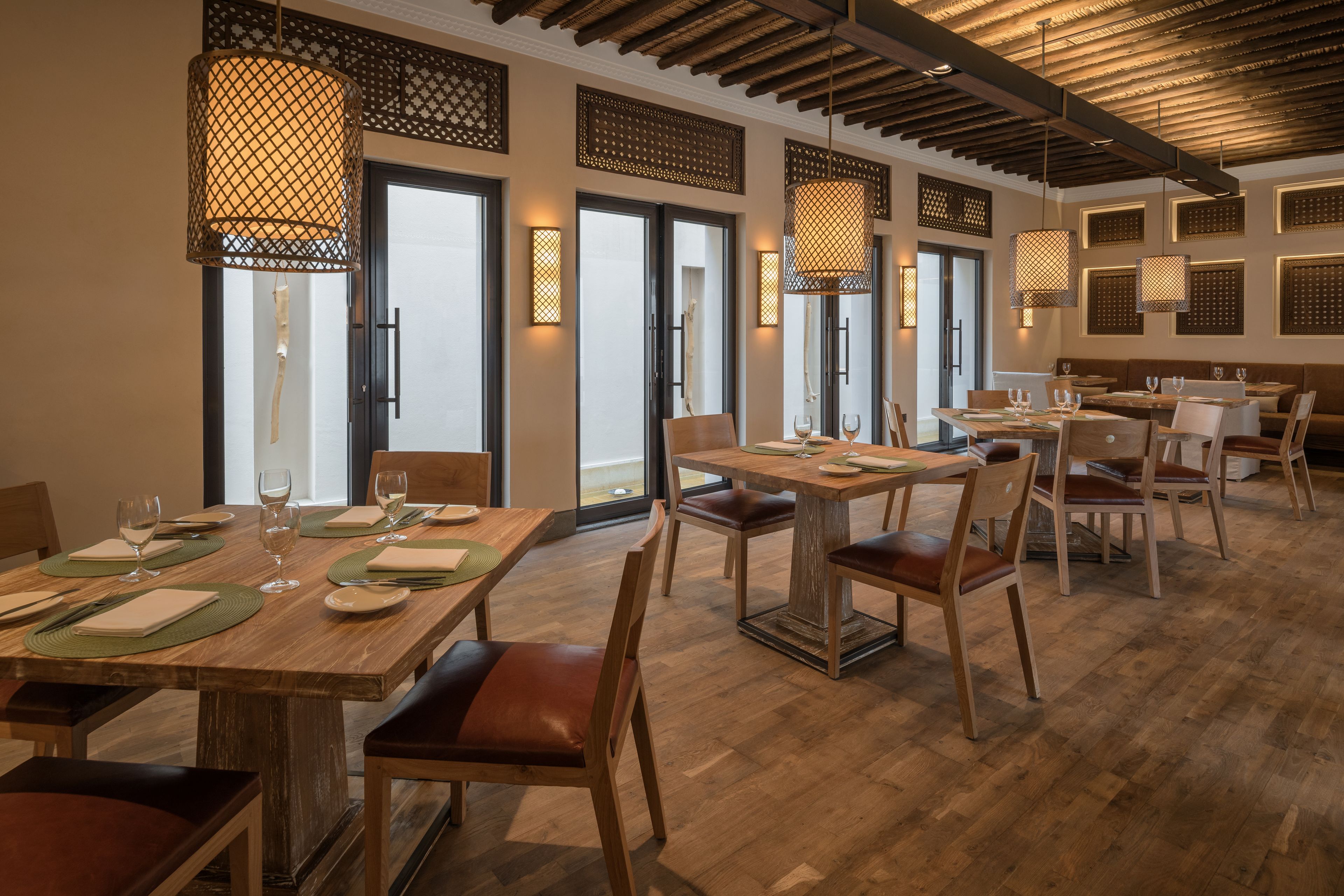 Um restaurante com mesas e cadeiras de madeira, iluminação pendente e grandes janelas.