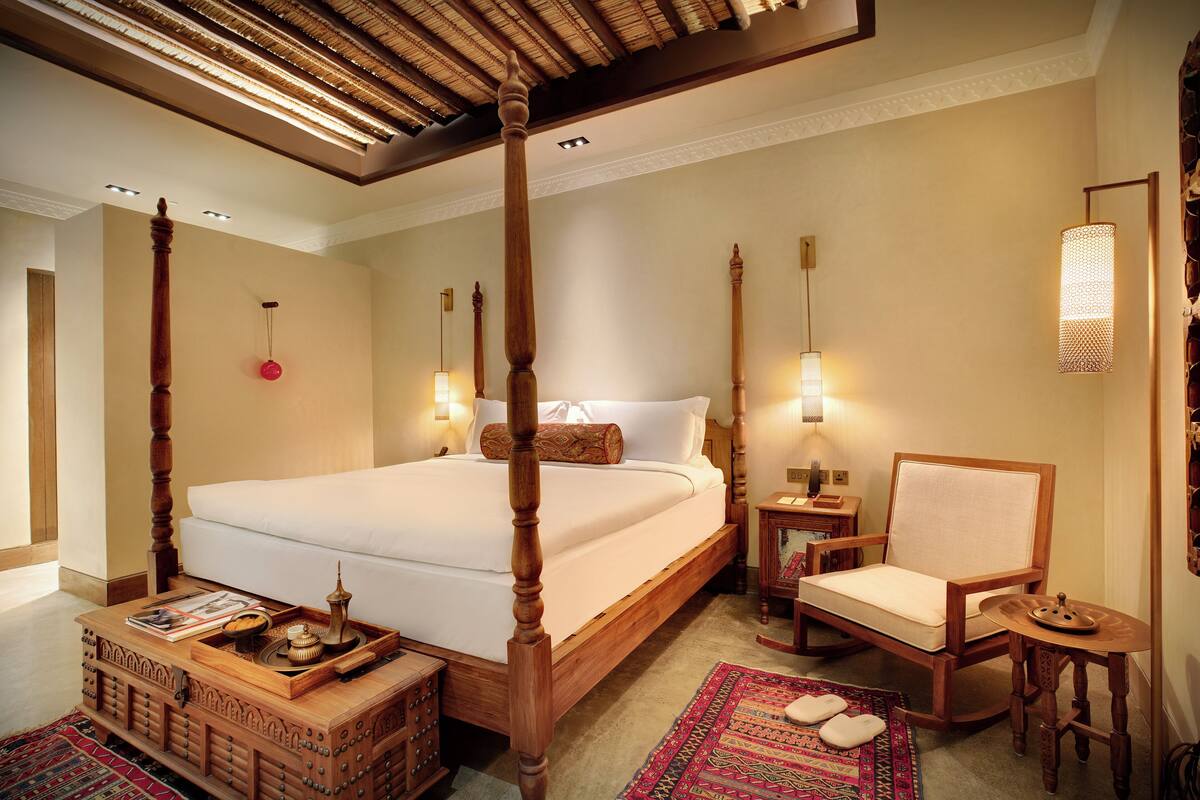 A four-poster bed with a wooden headboard, a wooden chair, a small table, and a decorative tray.