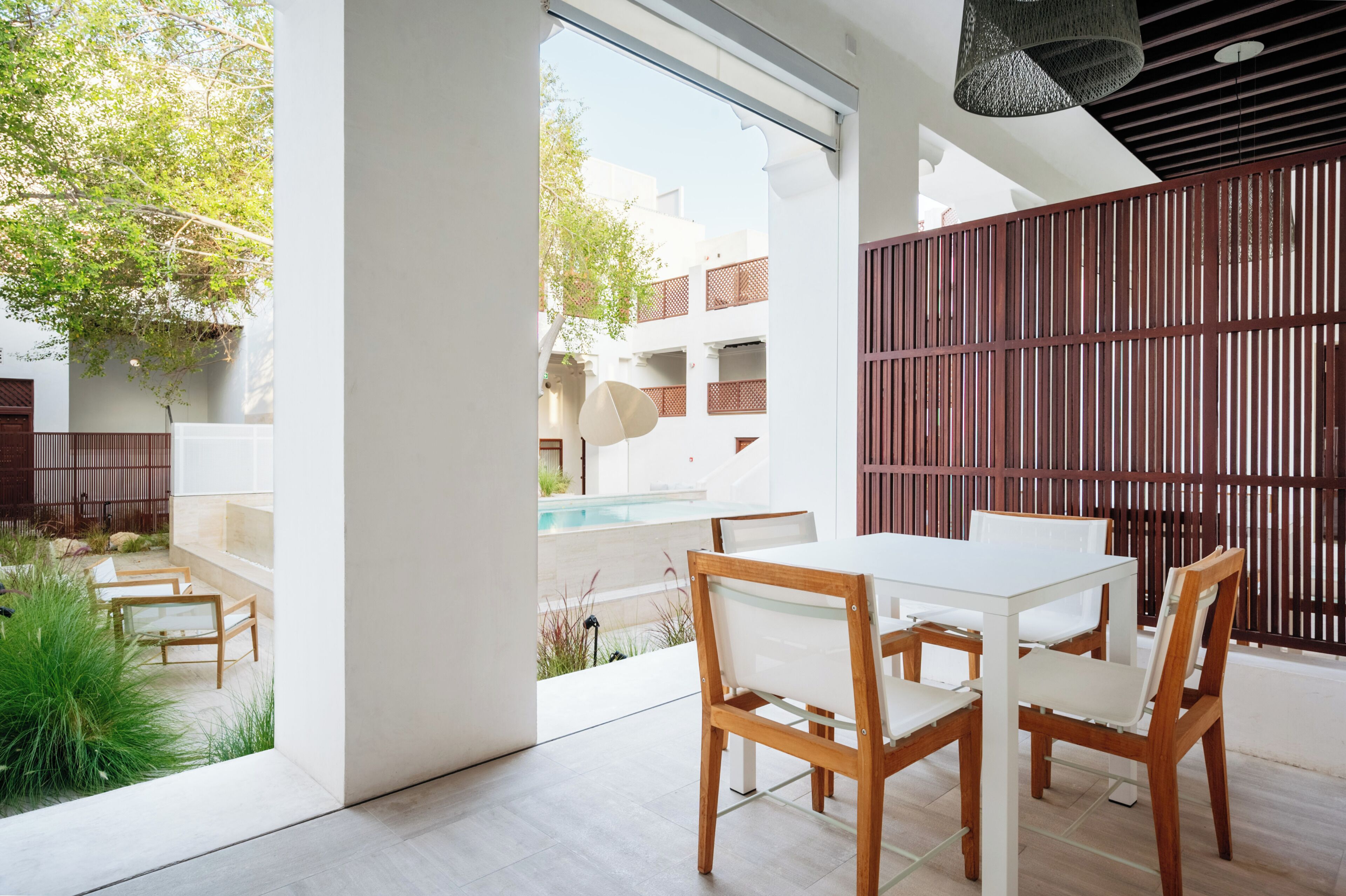 A modern outdoor dining area with a white table and chairs, a wooden fence, and a view of a building and greenery.