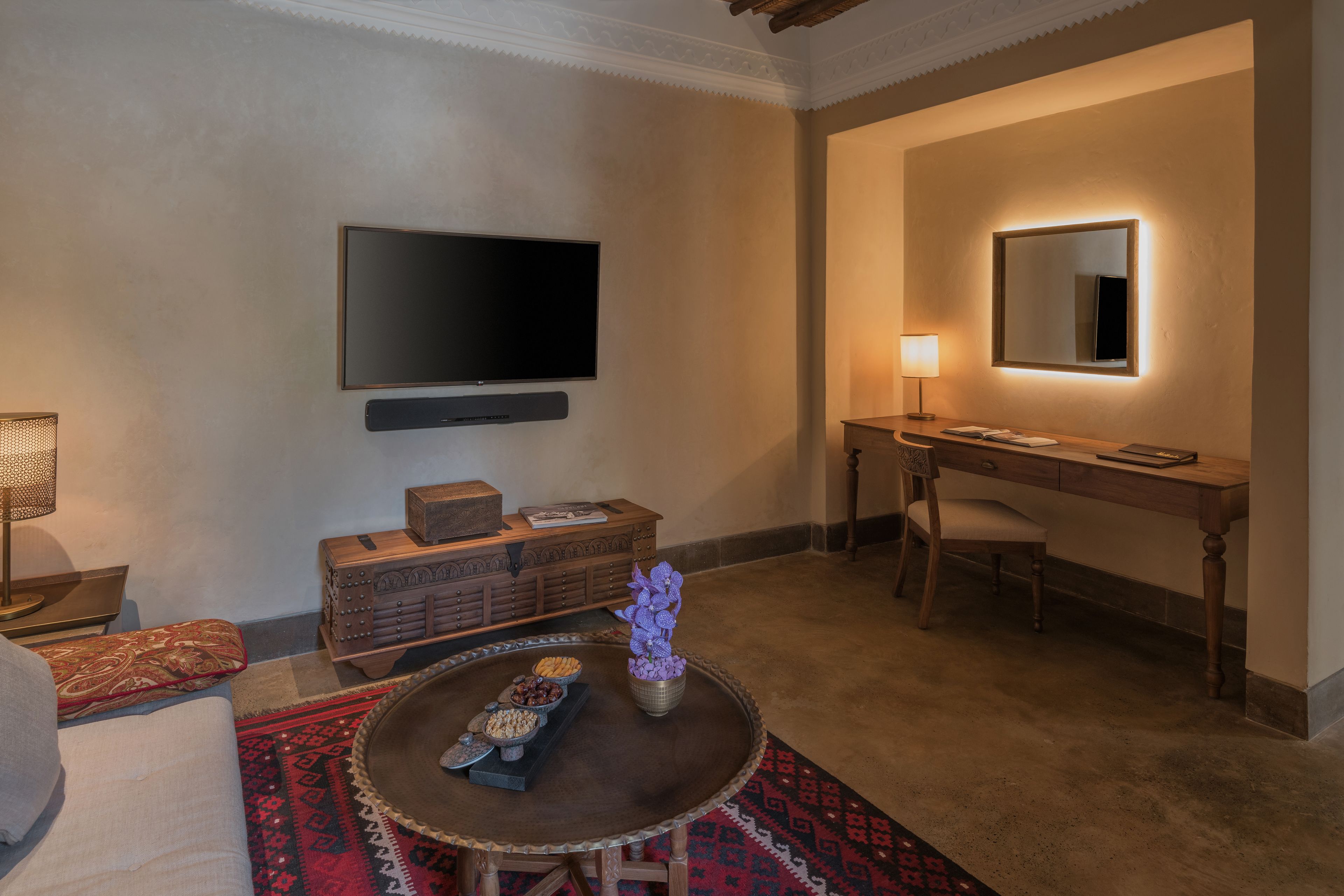 A modern living room with a flat-screen TV, a wooden desk with a lamp, a round coffee table with decorative items, and a patterned rug.