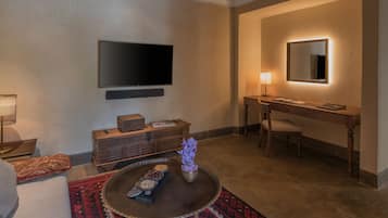 A modern living room with a flat-screen TV, a wooden desk with a lamp, a round coffee table with decorative items, and a patterned rug.
