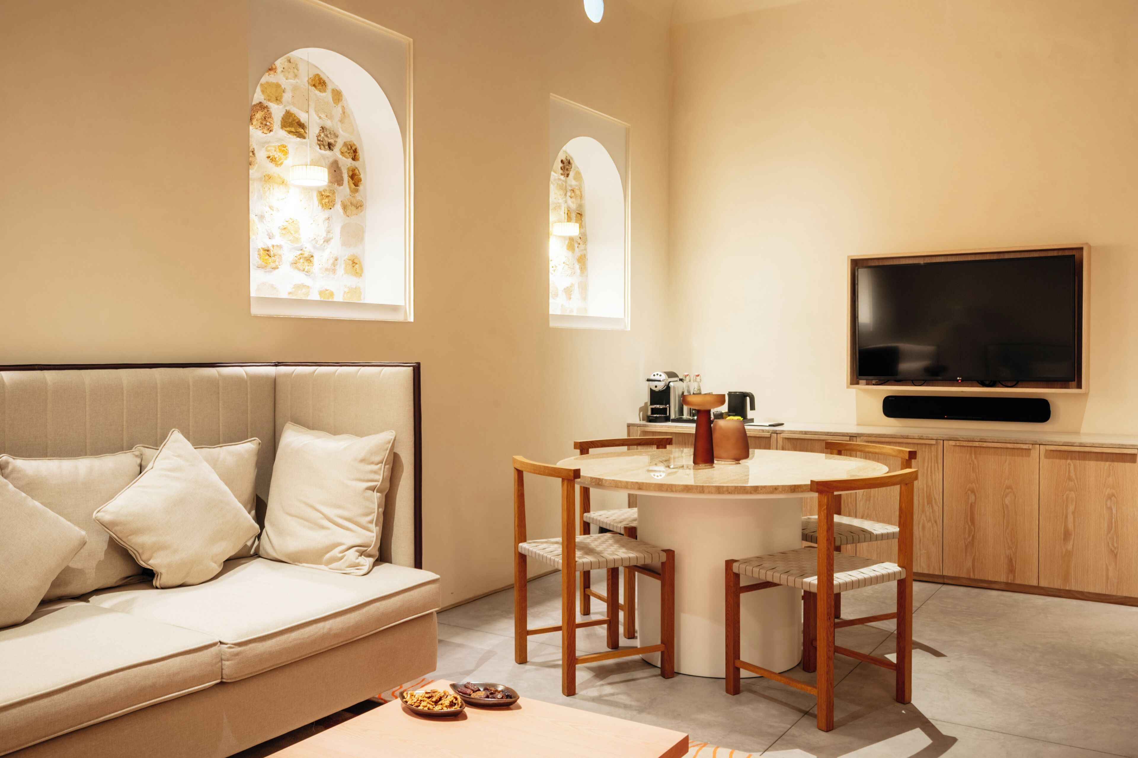 A modern living room with a sofa, a dining table, and chairs. There is a TV mounted on the wall and a window with a view.