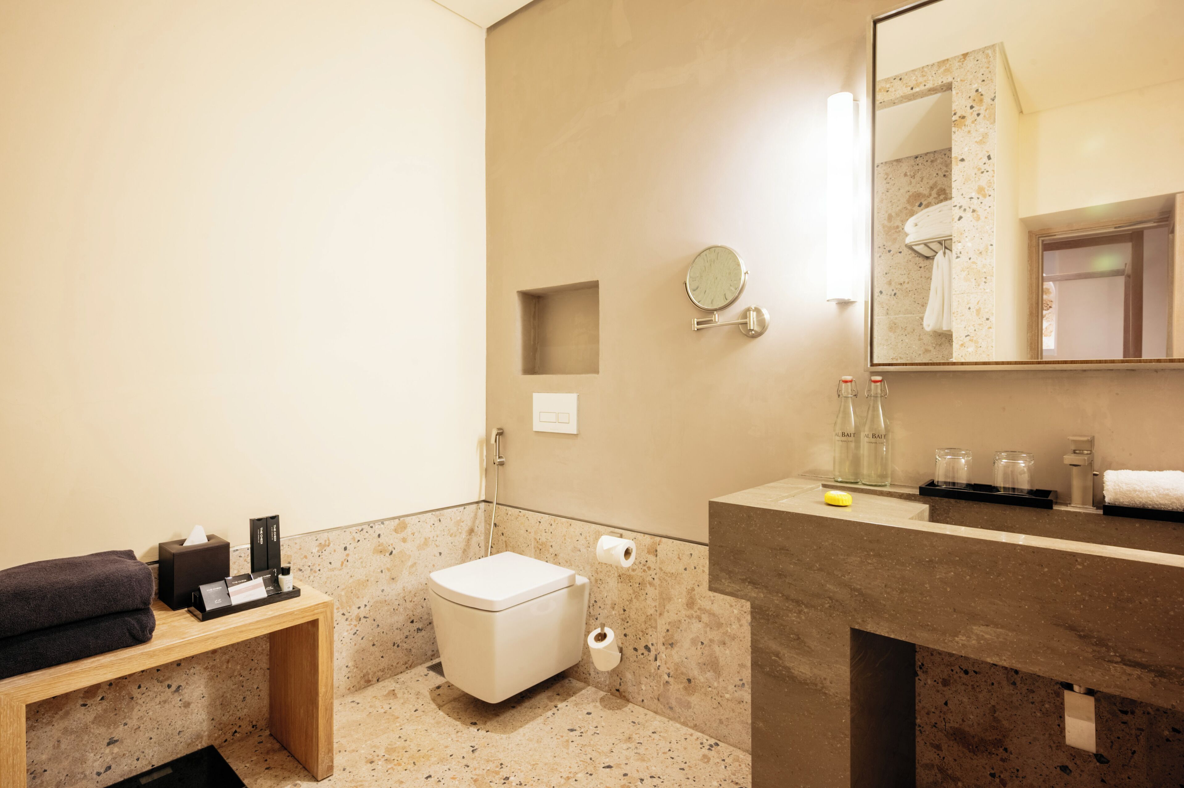 A modern bathroom with a toilet, sink, and mirror. There is a towel rack and a shelf with toiletries.