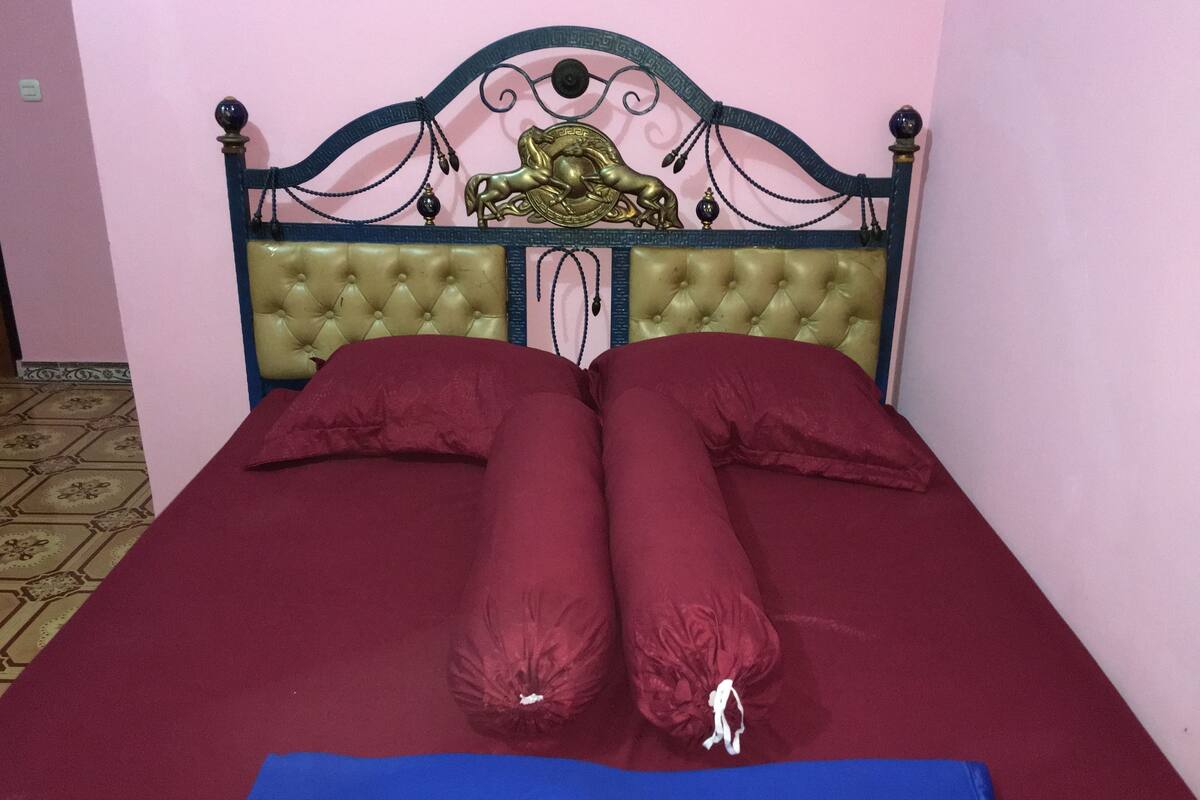 Una cama con cabecera verde, dos almohadas rojas y una manta azul.
