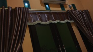 A window with dark curtains and a decorative trim, an air conditioning unit above, and a green-framed mirror to the left.