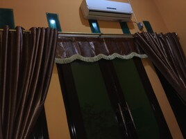 Una ventana con cortinas oscuras y detalles decorativos, una unidad de aire acondicionado arriba y un espejo con marco verde a la izquierda.