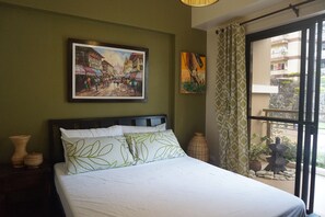 A neatly made bed with white linens and green leaf-patterned pillows, a framed painting of a street scene, a vase, and a view of a balcony with a stone lantern.