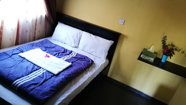 A bed with a blue and white bedspread, a white pillow, and a folded towel with a pink flower. There is a small shelf with a tissue box and a potted plant.