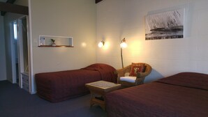 A room with two single beds, a small table with a cushion, a wicker chair, a wall-mounted shelf with a plant, and a painting. - Redwood Lodge Motel (Levin)