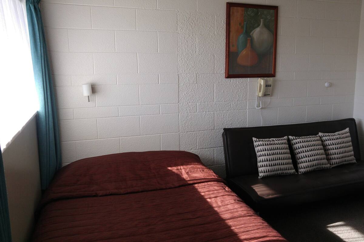 A room with a bed, a bench, a painting, and a telephone on the wall.