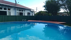 A swimming pool with a wooden deck and a bench, surrounded by a green fence and a house with a red roof. - Redwood Lodge Motel (Levin)