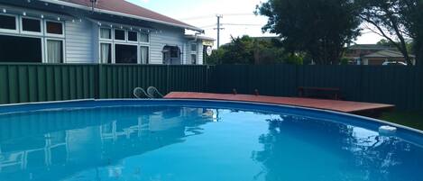 A swimming pool with a wooden deck and a bench, surrounded by a green fence and a house with a red roof.