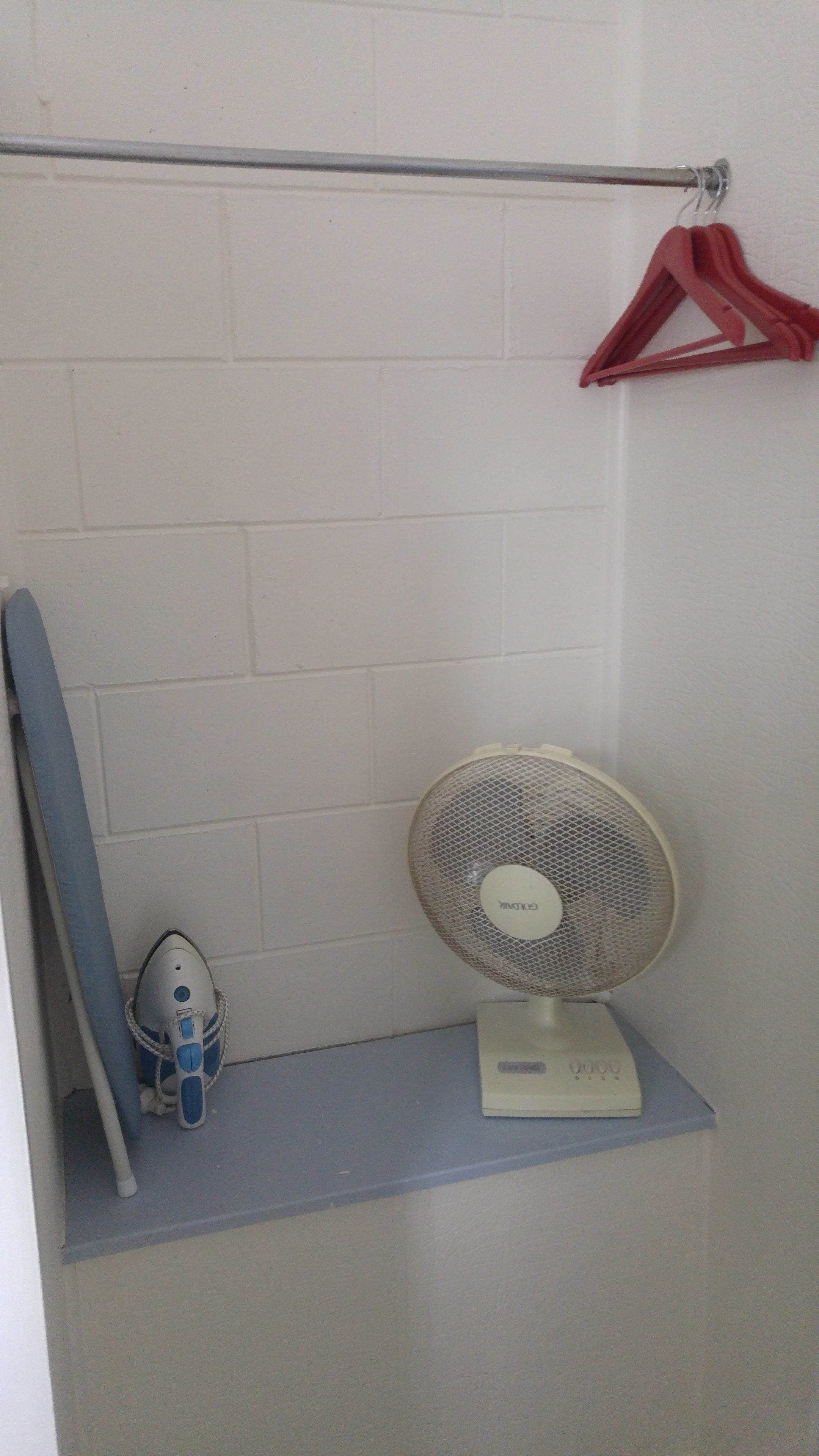 Ein Badezimmer mit weiß gefliesten Wänden, einem weißen Regal mit Ventilator und Bügelbrett sowie einem roten Kleiderbügel an einer Stange.