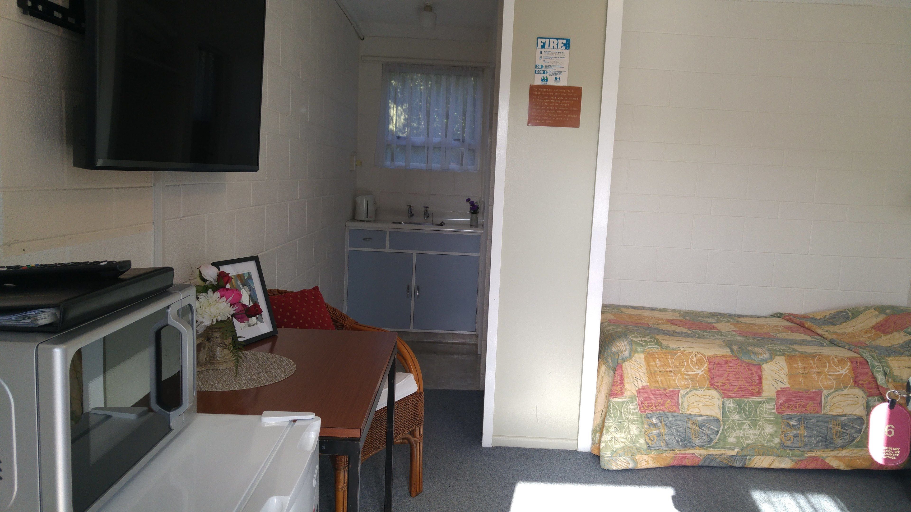 Een knus kamertje met een kitchenette, een bed met een kleurrijk sprei, een aan de muur bevestigde televisie, en een klein tafeltje met een fotolijstje en bloemen.