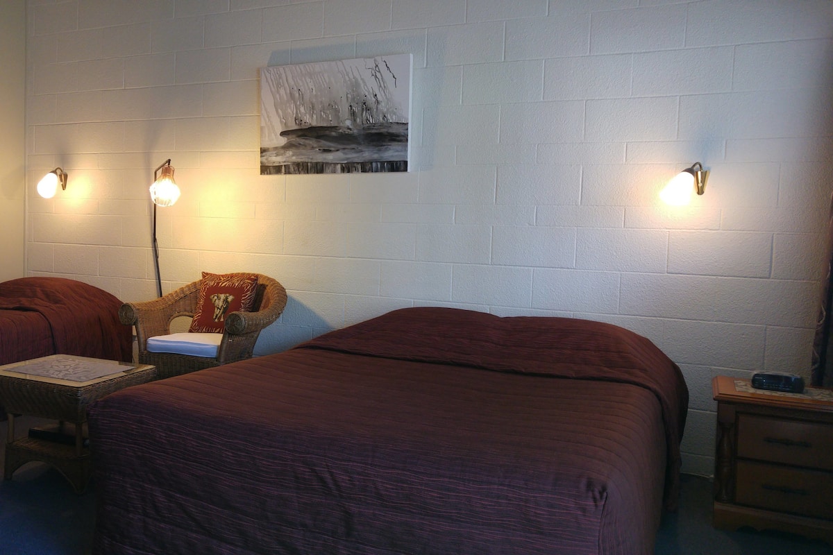 A room with two beds, a small table, a wicker chair, and a wall-mounted painting.