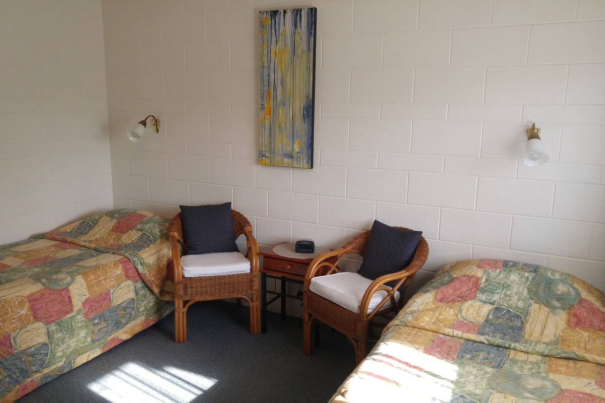 A room with two single beds, a small table, two wicker chairs, and a wall-mounted light fixture.