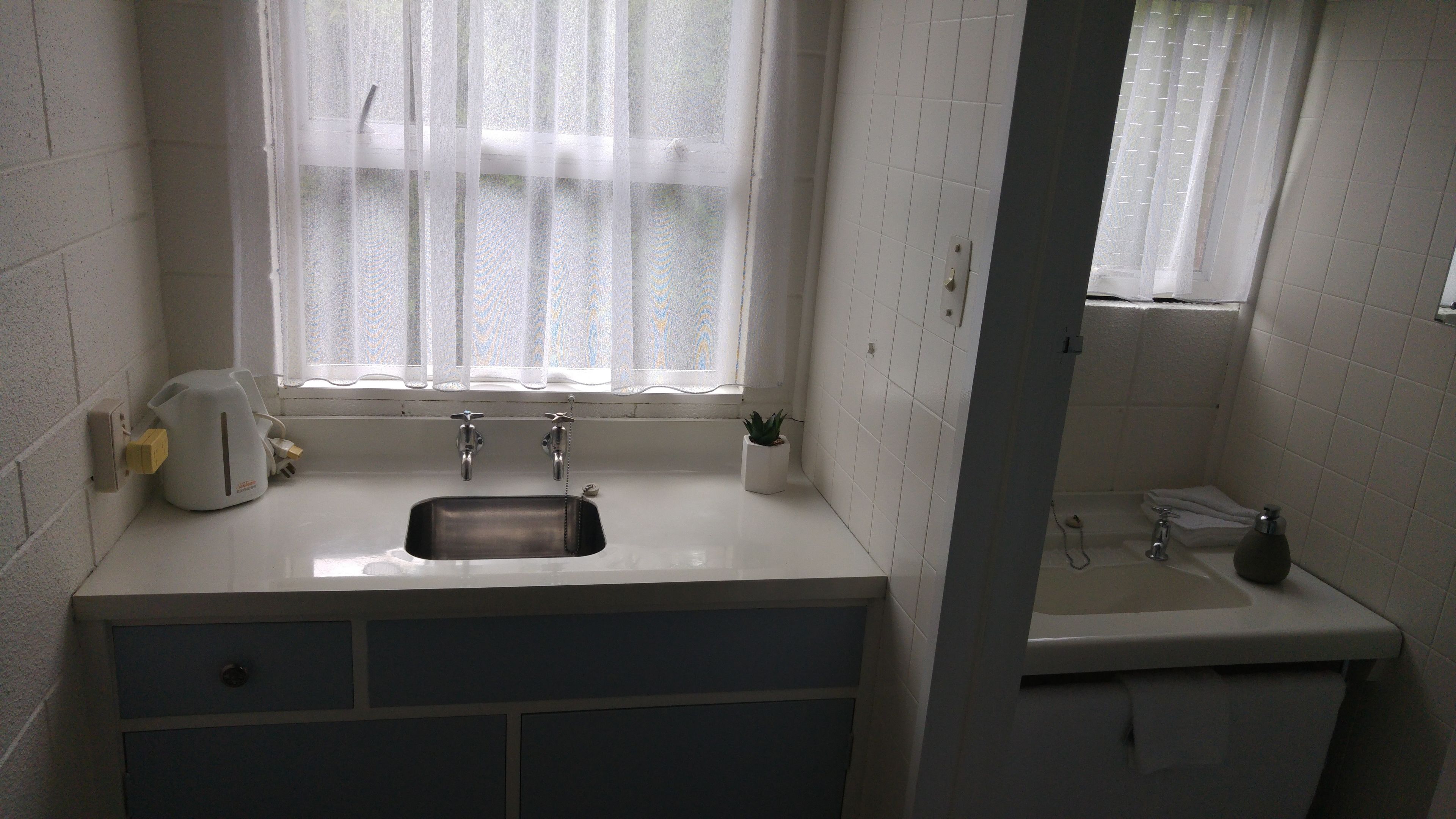 Un baño pequeño y limpio con un lavamanos blanco, un espejo y una ventana con cortinas translúcidas.