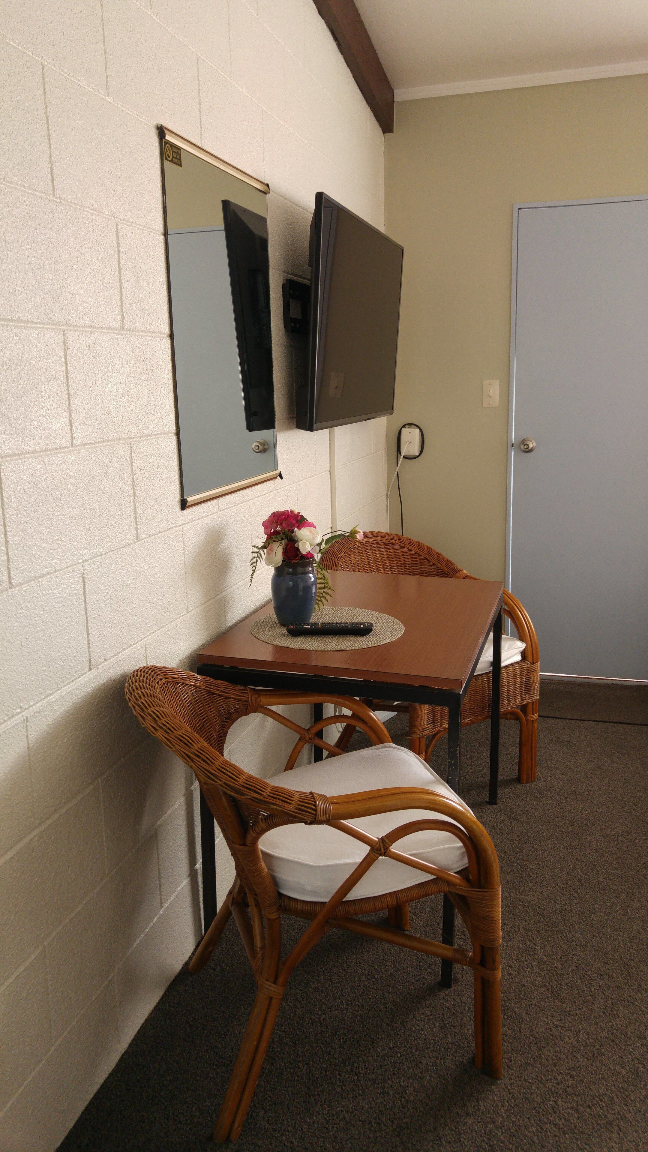 A room with a television mounted on the wall, a wicker chair, a small table with a vase of flowers, and a door.