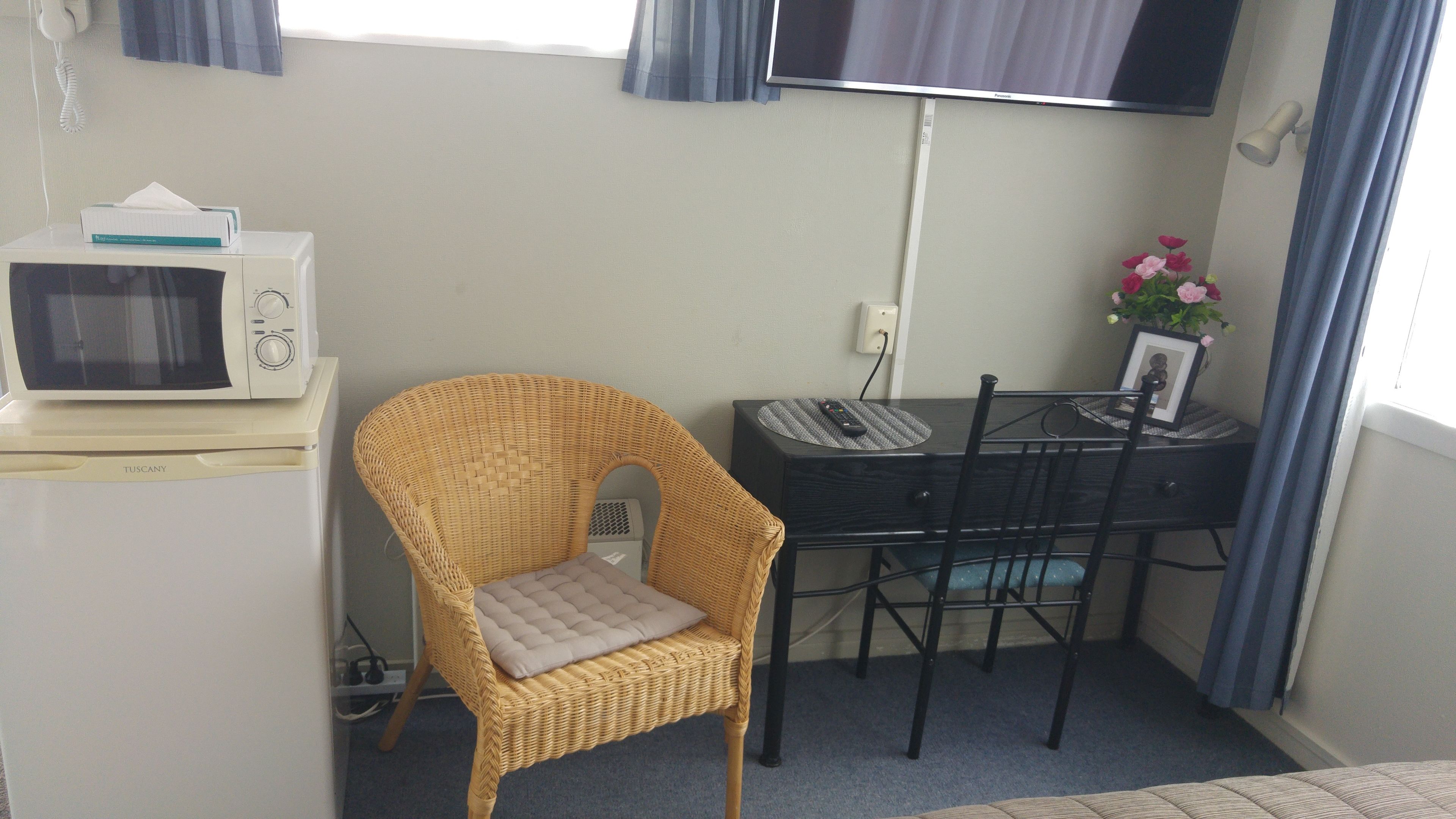 A compact room with a white microwave, a wicker chair with a cushion, a small table with a remote control, and a TV mounted on the wall.