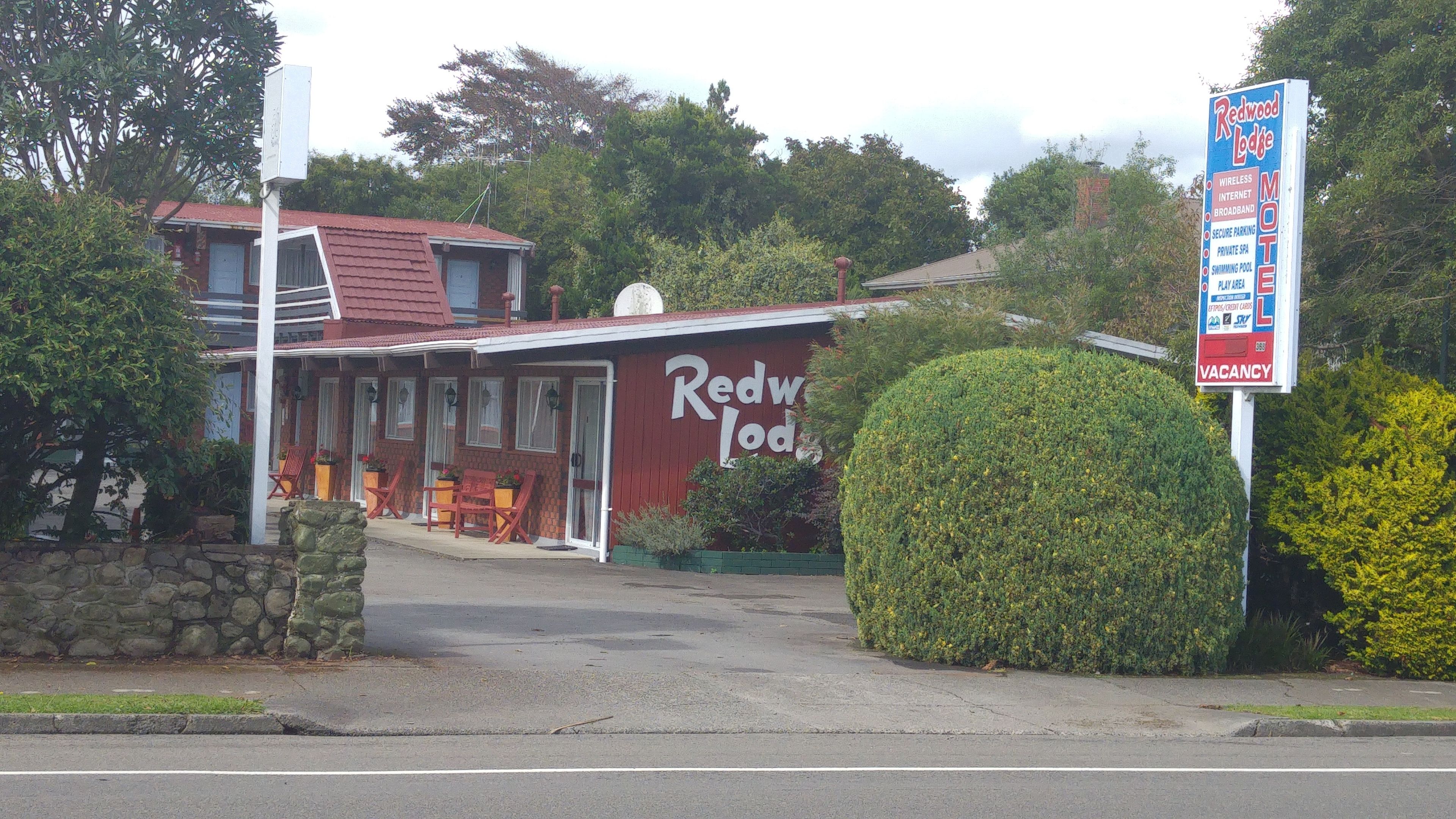 A Redwood Lodge motel with a red exterior, a covered outdoor seating area, and a signboard listing amenities like wireless internet and a swimming pool.