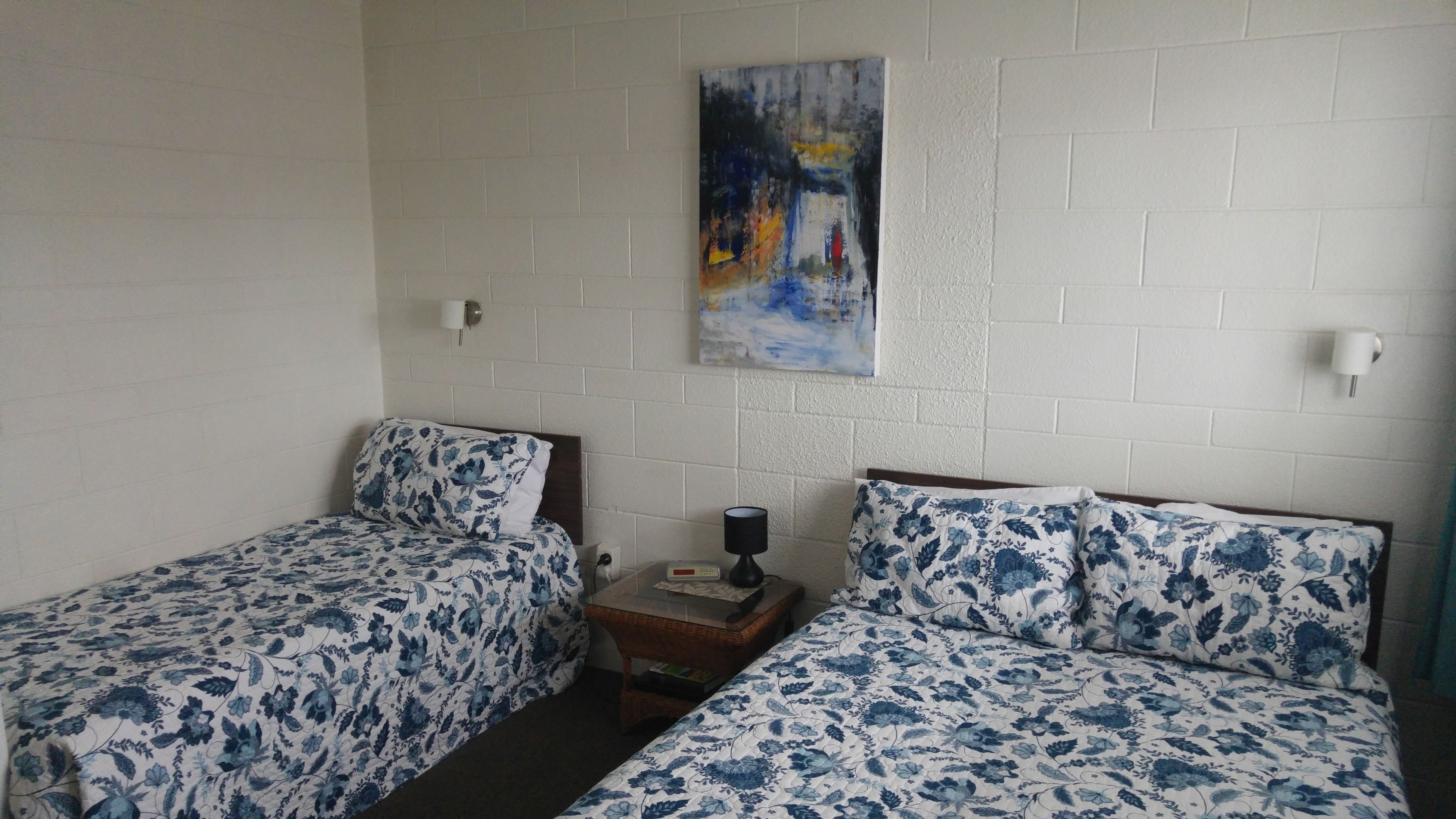 A room with two single beds, each with floral-patterned bedding, a small wooden nightstand with a lamp, and a painting on the wall.