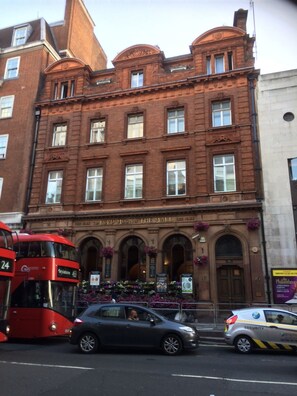 A red double-decker bus parked in front of a building with a classic architectural style, featuring arched windows and a central entrance. - Tsq Whitehall (London)