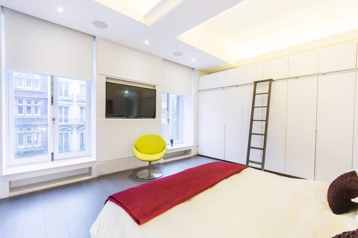 Une chambre moderne avec un lit, un plaid rouge, un fauteuil jaune, une télévision et de grandes fenêtres.