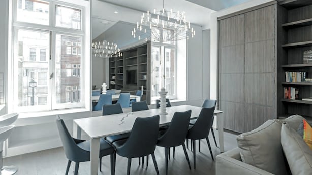 A modern dining area with a white table and dark chairs, large windows, and a bookshelf. - Tsq Whitehall (London)