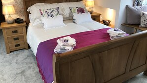 A neatly made bed with a wooden headboard, two bedside tables with lamps, a floral-patterned wall, and a small window with curtains. - Lon Y Traeth Bed & Breakfast (Pentraeth)