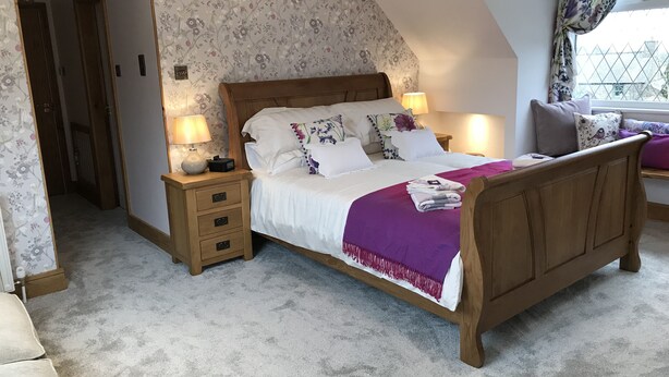 A bedroom with a wooden bed, two bedside tables with lamps, a nightstand, a window with curtains, and a floral-patterned wall. - Lon Y Traeth Bed & Breakfast (Pentraeth)