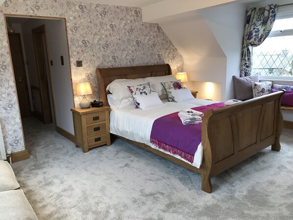 A bedroom with a wooden bed, two bedside tables with lamps, a nightstand, a window with curtains, and a floral-patterned wall. - Lon Y Traeth Bed & Breakfast (Pentraeth)