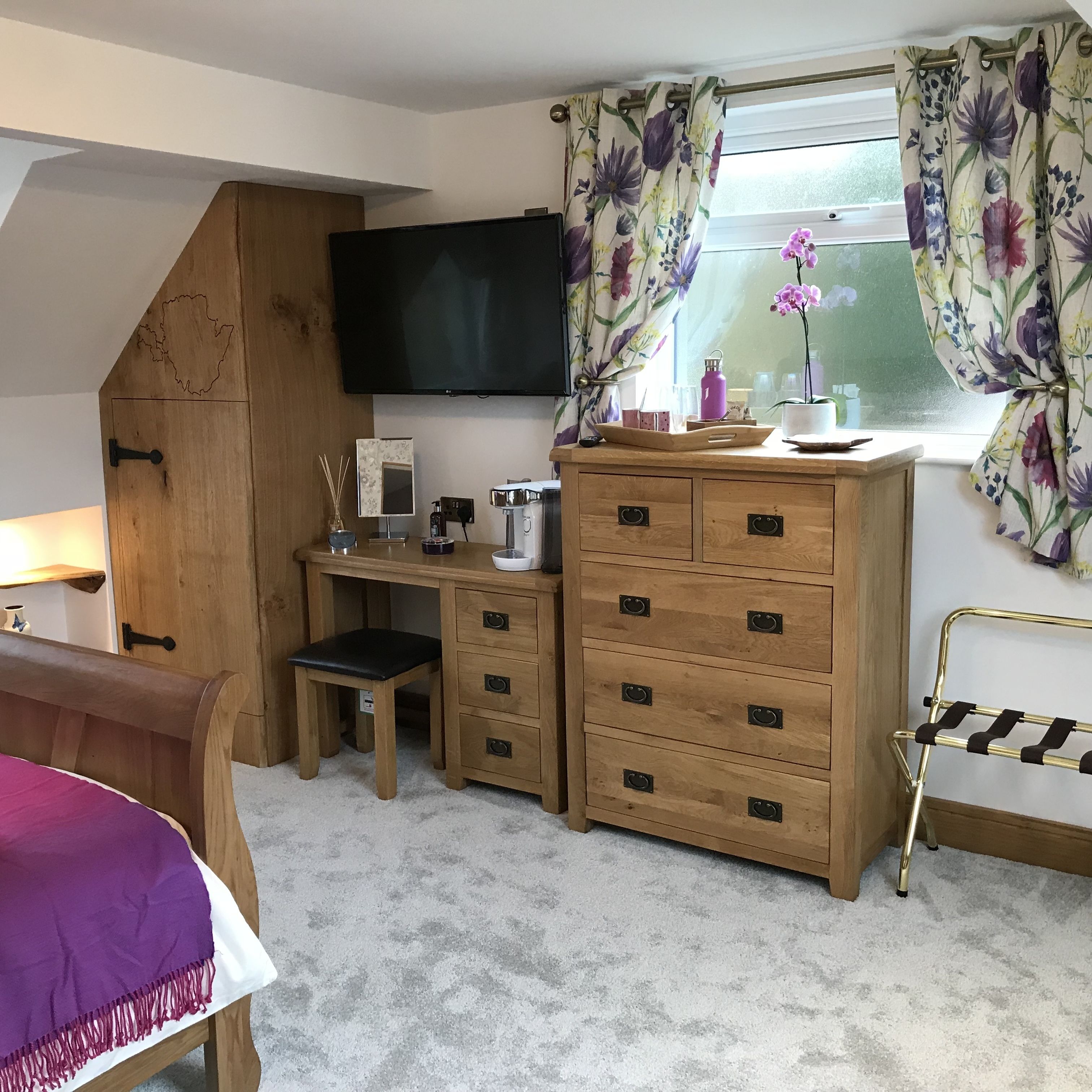 Un dormitorio con escritorio de madera, cama con colcha morada, cómoda con varios cajones, televisor montado en la pared y ventana con cortinas florales.