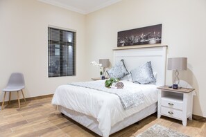 Quarto com cama, mesa de cabeceira, cadeira, janela com persianas e pintura de paisagem urbana na parede.