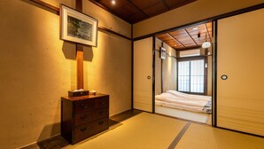 Perinteinen japanilaistyylinen huone, jossa on tatamimatto, puinen lipasto laatikoineen, seinällä oleva kehystetty kuva ja liukuovi, joka johtaa valkoisilla vuodevaatteilla varustettuun sänkyyn.