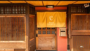 Entrada de um edifício tradicional japonês, com portas de correr de madeira e uma faixa amarela com o nome "Kumo Machiya".