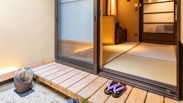 Una habitación japonesa tradicional con suelo de tatami, una puerta corredera y un pequeño jardín con una roca, una planta y un par de zapatillas.