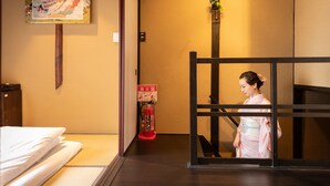 Una habitación japonesa tradicional con suelo de tatami, una cama con ropa de cama blanca, un extintor y una persona con un kimono rosa.