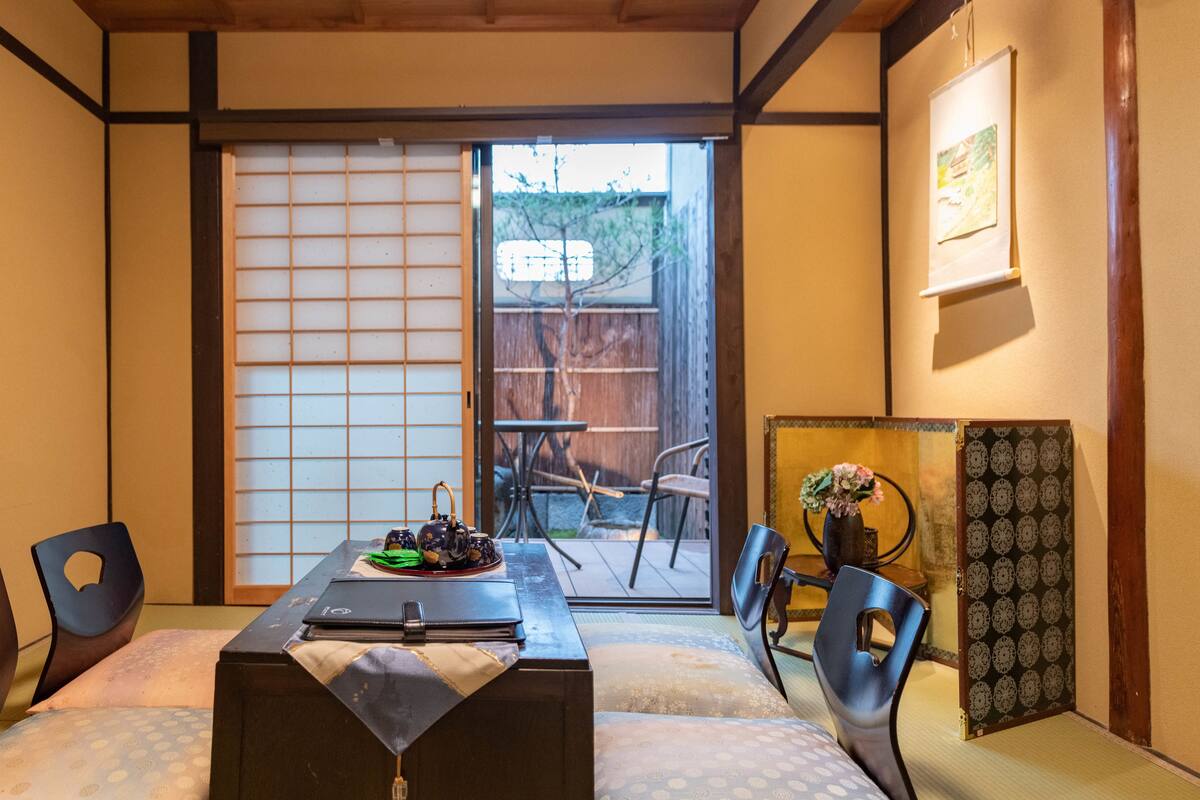 A traditional Japanese room with tatami flooring, a sliding shoji screen, a low table with a tea set, and a view of a garden through the screen.