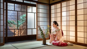 Um quarto japonês tradicional com tatames, painéis shoji e uma pessoa vestindo quimono segurando um arranjo de flores.