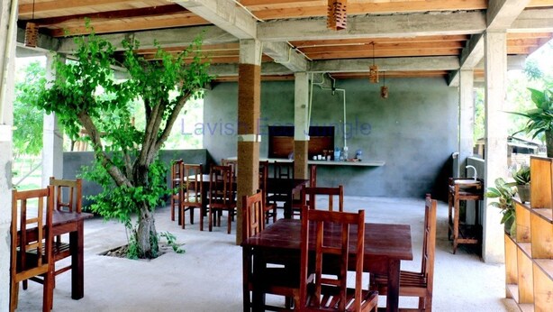 An indoor dining area with wooden tables and chairs, a large tree, and a concrete wall with a grill.