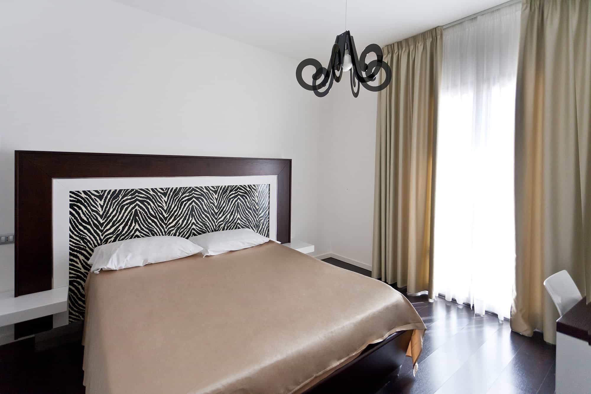 A hotel room with a zebra-patterned headboard, a bed with a beige cover, a modern light fixture, and a window with curtains.