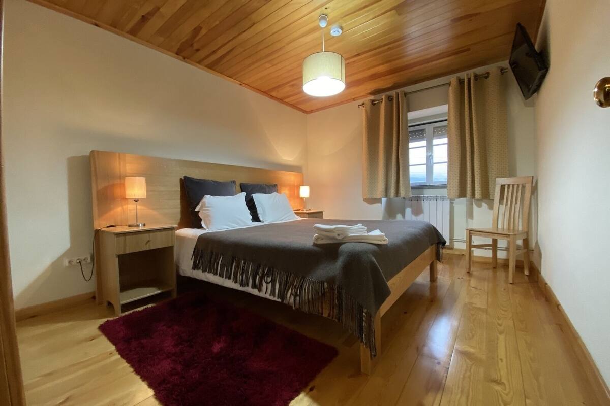 A bedroom with a wooden ceiling, a bed with a dark bedspread, two bedside lamps, a red rug, a wooden chair, and a window with curtains.