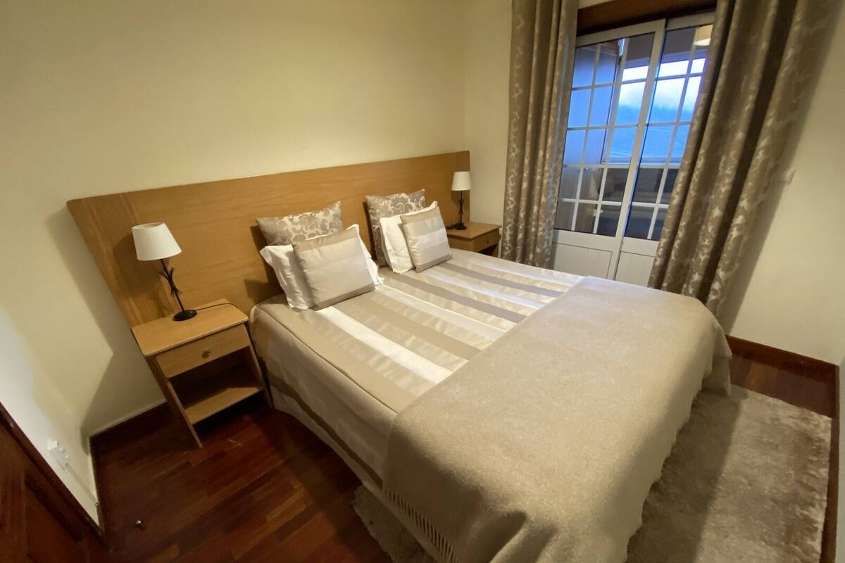 A neatly arranged bedroom with a wooden headboard, a large bed with striped bedding, two bedside tables with lamps, and a view of the sky through a window with curtains.