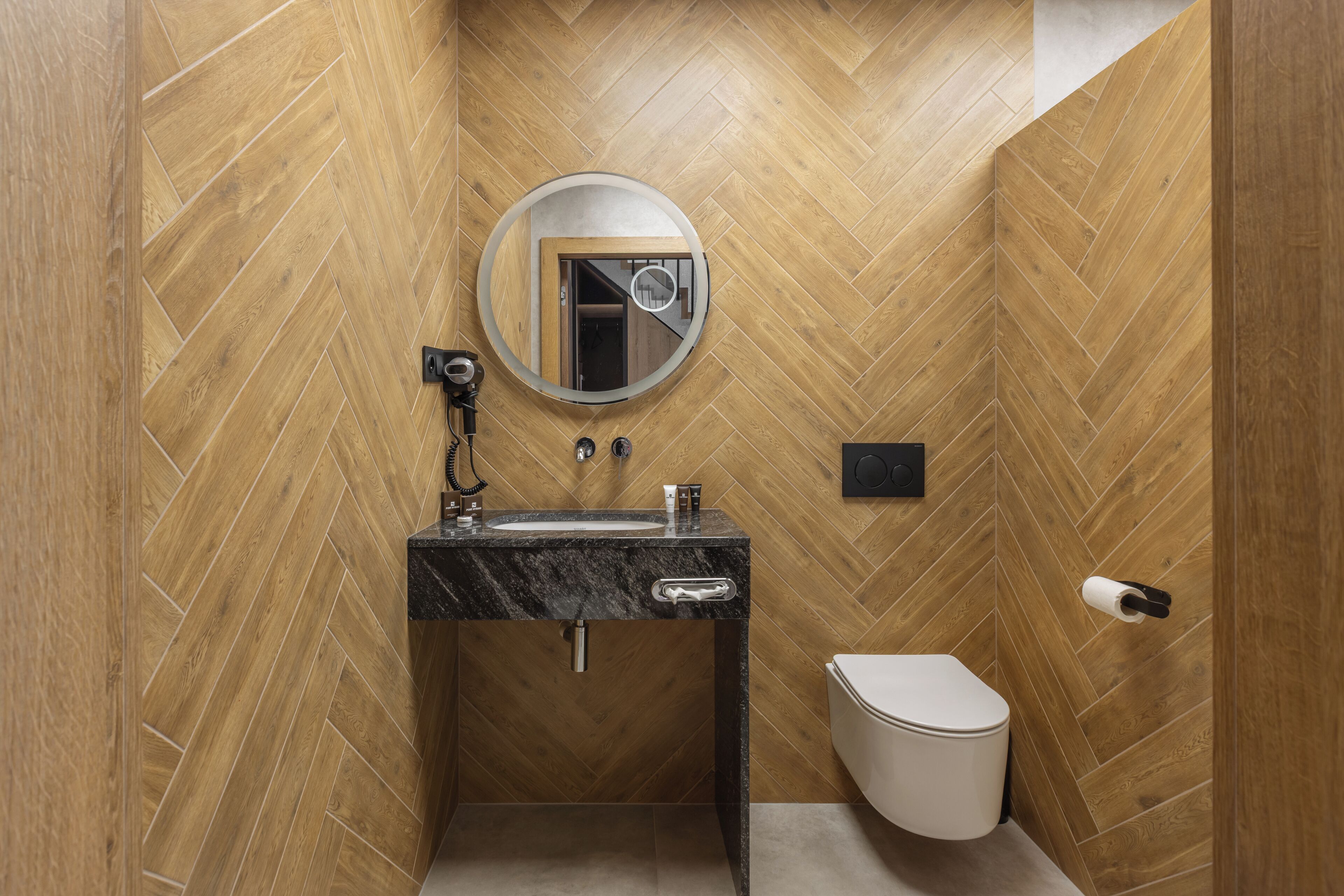 A bathroom with a herringbone wood floor, a round mirror, a black marble sink, a toilet, and a towel dispenser.