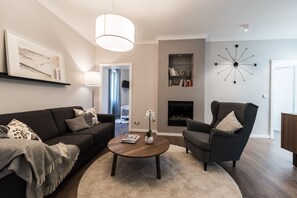 Een moderne woonkamer met een donkere bank, een ronde salontafel en een grijze fauteuil. Er is een open haard en een klok aan de muur.