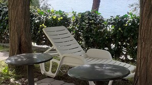 A seating area with two white plastic lounge chairs and two small round tables, surrounded by trees and a view of a body of water.